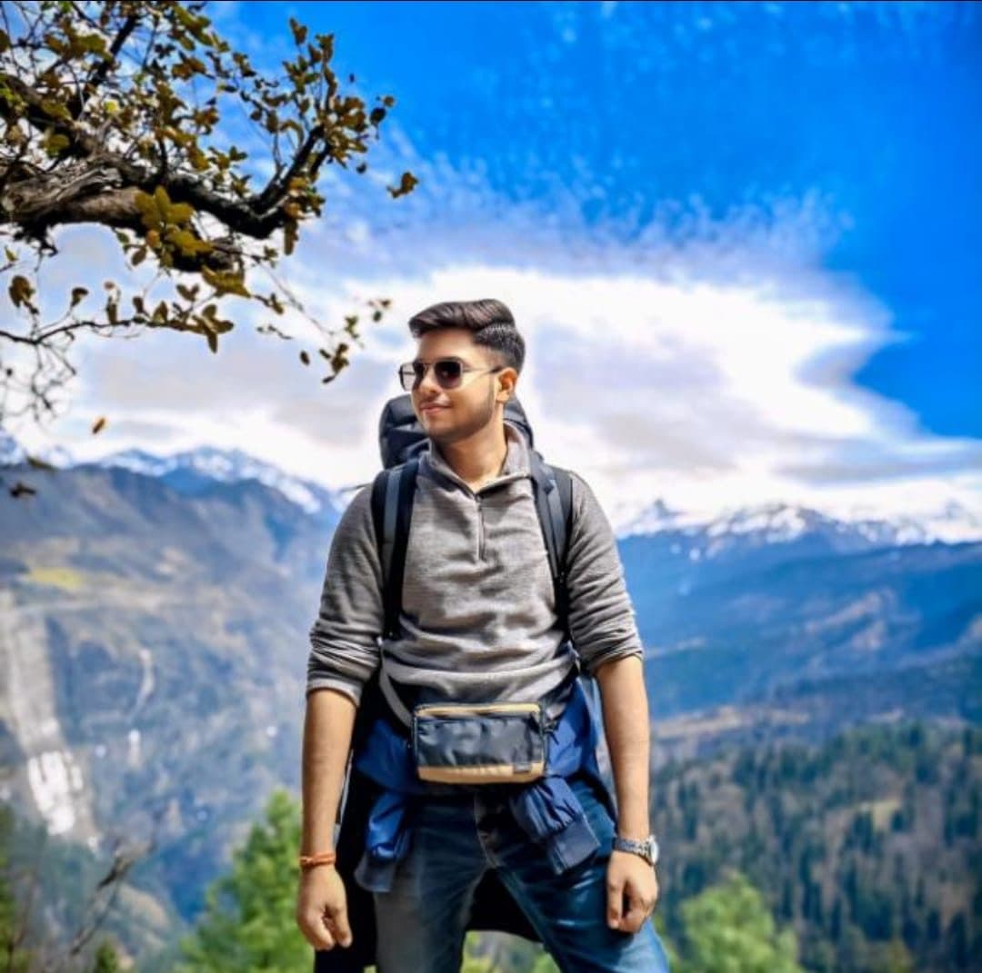 Harshit standing on a mountain trail with a backpack, snow-capped peaks behind him