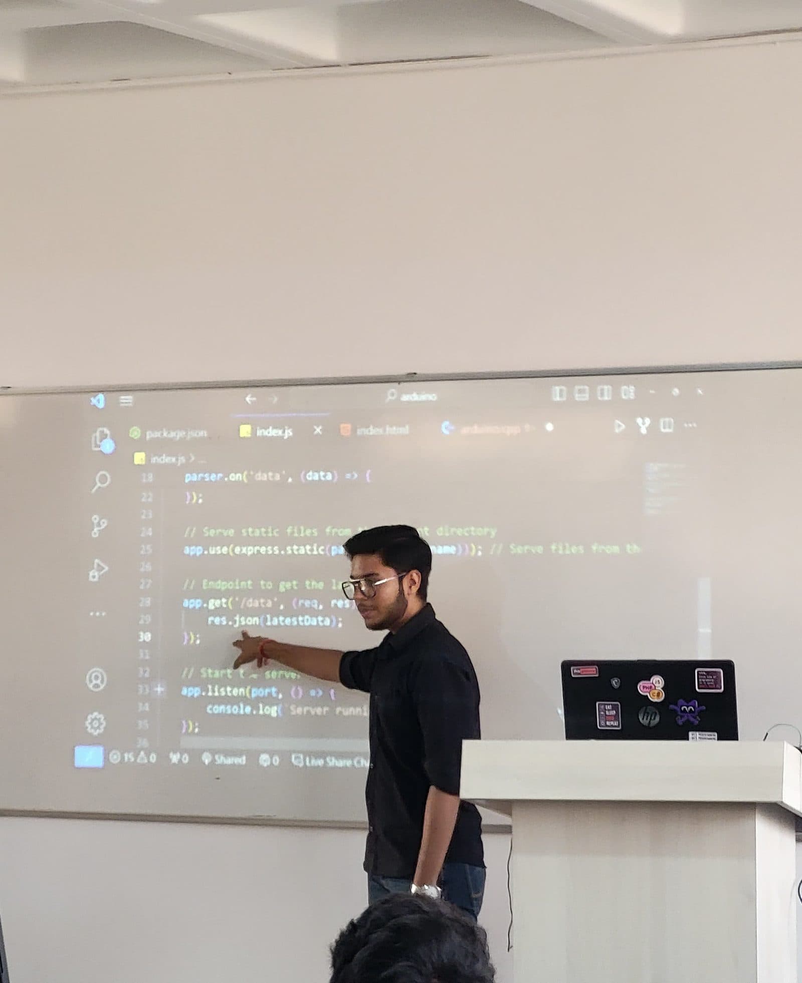 Harshit pointing at Arduino and Node.js code projected on a whiteboard during a workshop