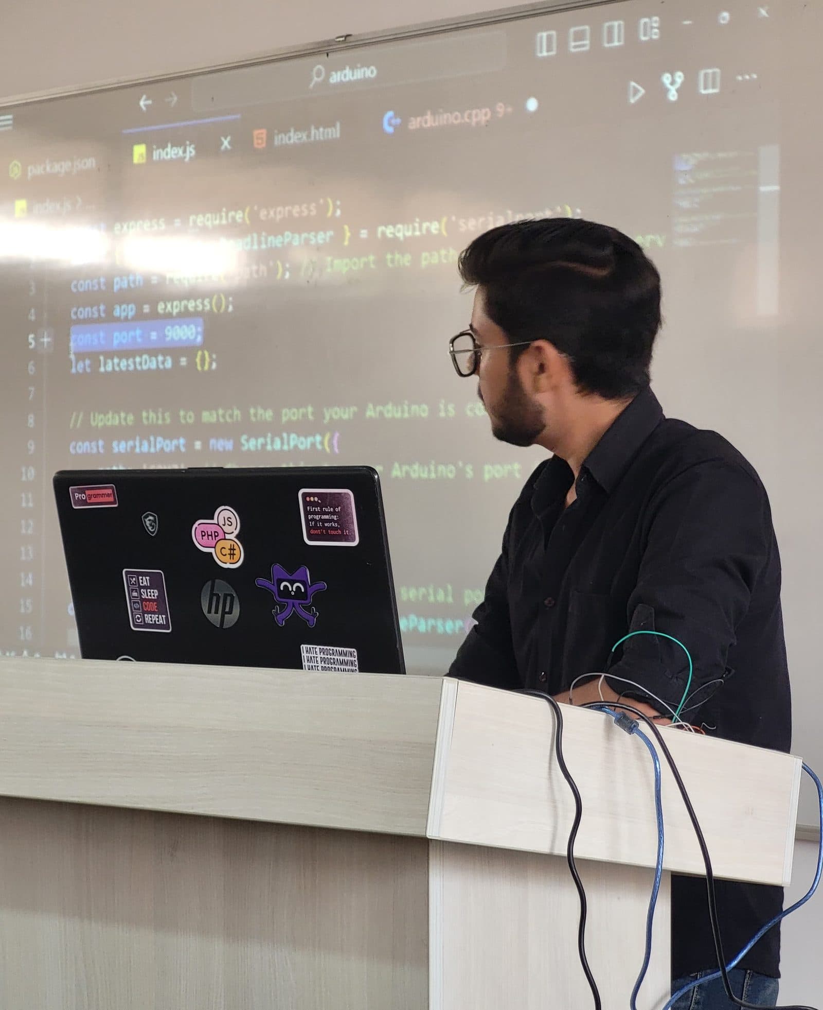 Harshit at a podium with a sticker-covered laptop, projected code behind him